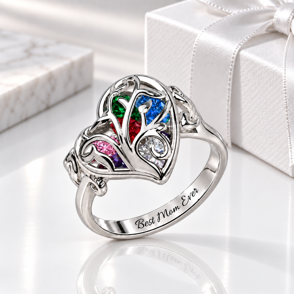 Silver Family Tree Heart Cage Ring with colorful heart-shaped birthstones and “Best Mom Ever” engraving, displayed on a glossy white surface beside a white gift box.