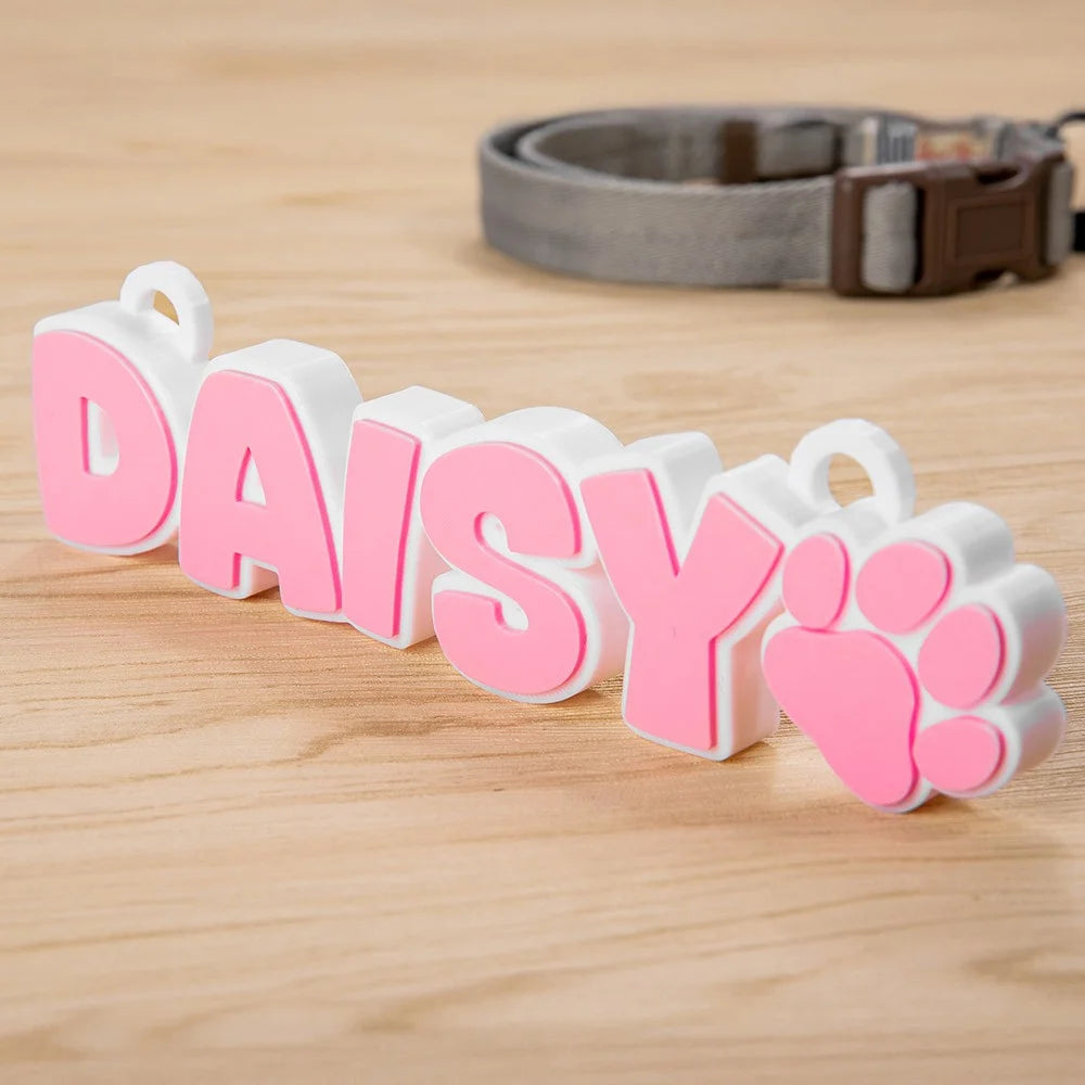 Pink and white custom pet name tag 'DAISY' with paw print, personalized dog ID tag on wood surface