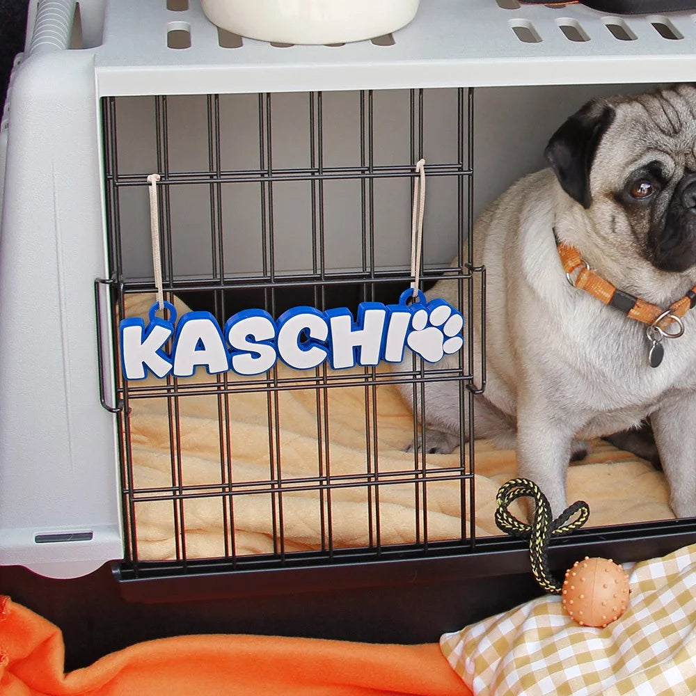 Personalized dog crate tag with name KASCHI and paw print, featuring a pug inside a cozy crate.