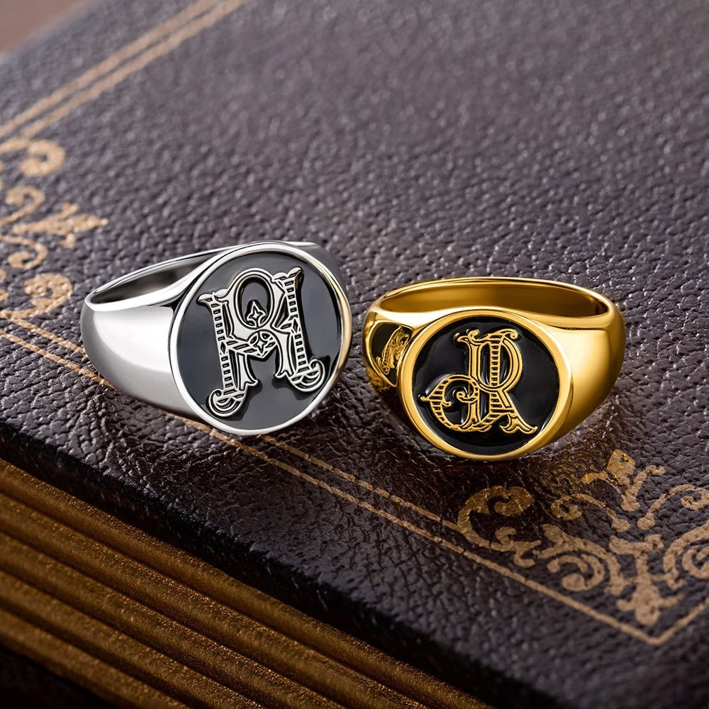 Silver and gold tone custom monogram signet rings with intertwined initials displayed on a decorative book.
