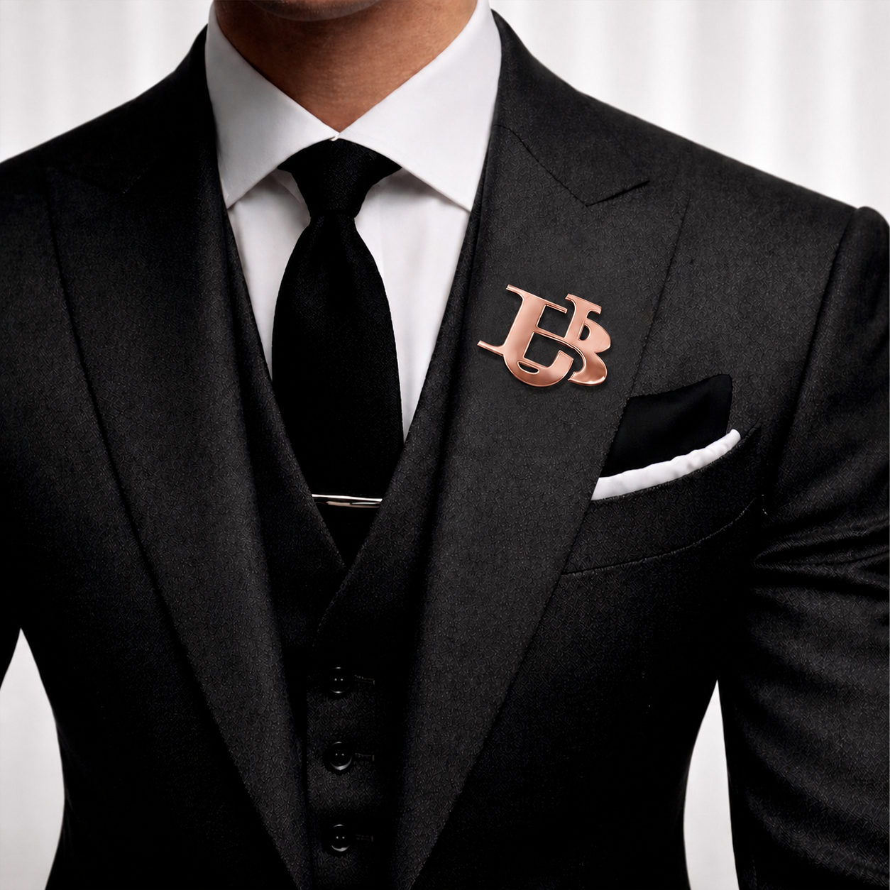 Rose gold interlocking initials lapel pin worn on a black suit with a white shirt and black tie.