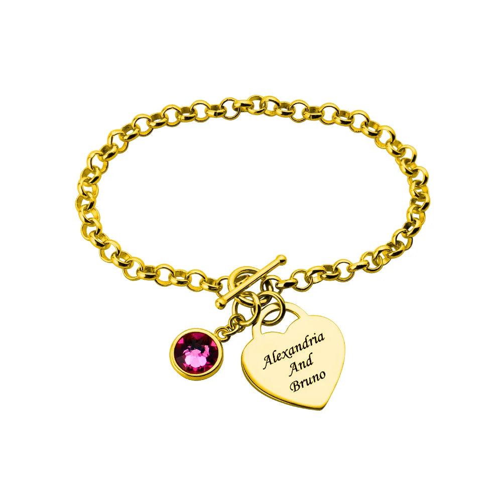 Gold-plated personalized heart charm bracelet with a pink birthstone and engraved names "Alexandria and Bruno," featuring a secure toggle clasp and elegant link design.