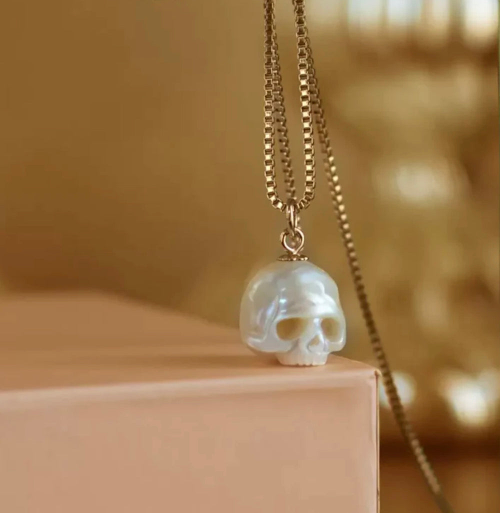 Opalescent skull pendant on a gold box chain necklace, resting on a soft peach-colored edge with a blurred golden backdrop.
