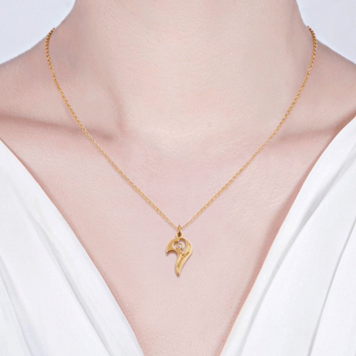 A gold heart-shaped pendant with a gemstone on a delicate gold chain, worn around the neck of a person in a white top.