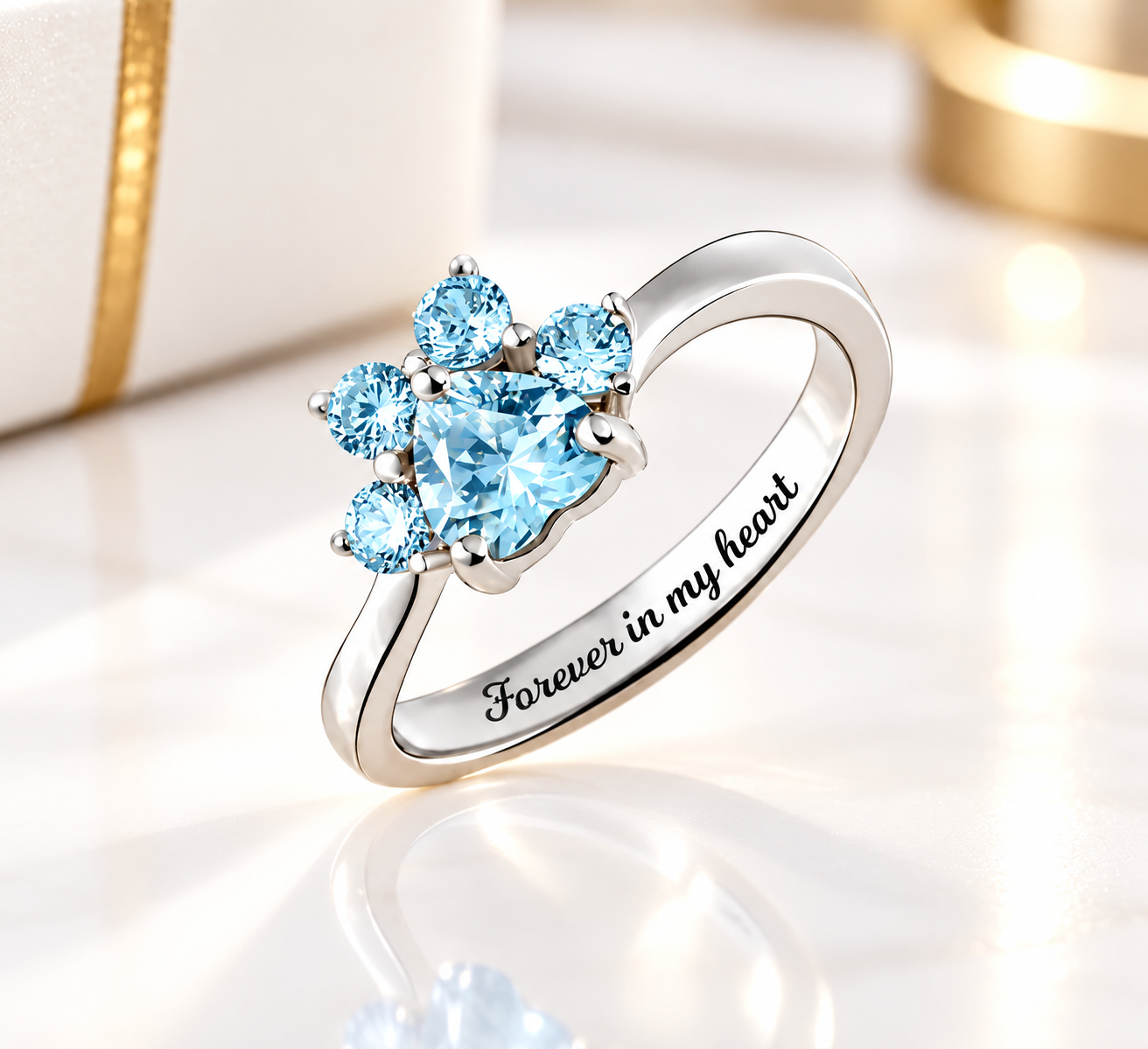 Aquamarine blue heart-shaped pet paw print ring in sterling silver, featuring sparkling blue gemstones and “Forever in my heart” engraving, displayed on an elegant white and gold background.