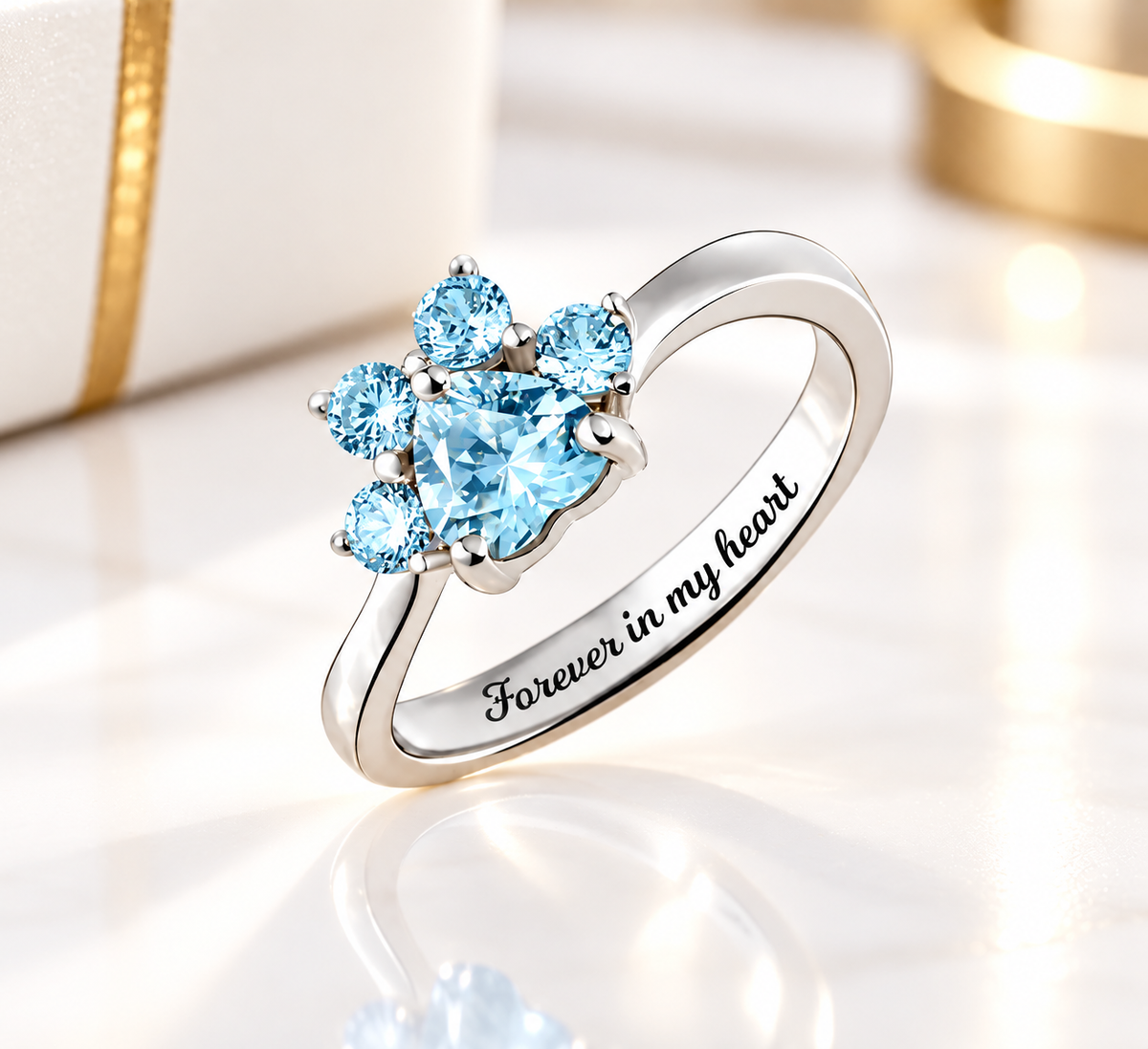 Aquamarine blue heart-shaped pet paw print ring in sterling silver, featuring sparkling blue gemstones and “Forever in my heart” engraving, displayed on an elegant white and gold background.