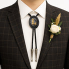 Personalized bolo tie with gold initial 'L', worn with plaid suit and white rose boutonniere for formal occasions.