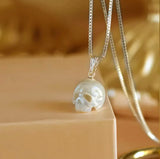 A glowing skull pendant dangles from a fine silver necklace, placed gently on a beige surface with a golden hue in the background.