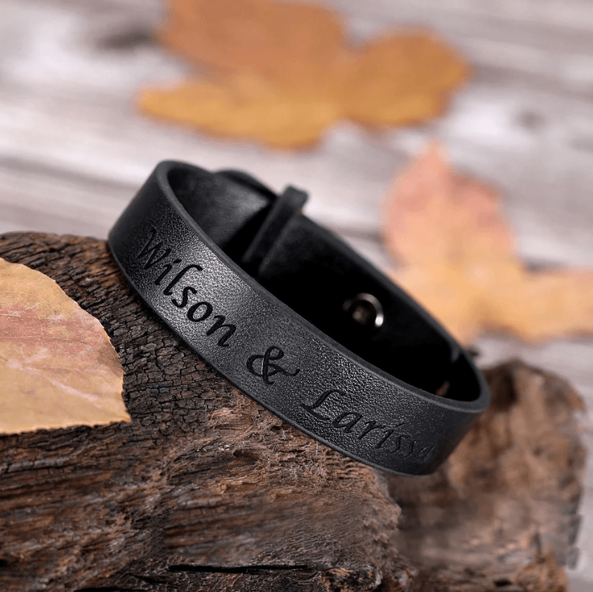 Custom engraved black leather bracelet displayed on a rustic wooden log with autumn leaves in the background.