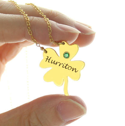 A hand holding a gold necklace with a four-leaf clover pendant inscribed with "Huririton" and a green gemstone.