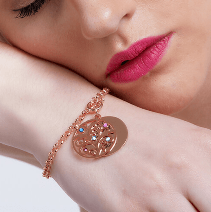 A woman wearing a rose gold charm bracelet with a tree pendant adorned with colored gemstones and a round disc, resting her head on her arm, with pink lips.