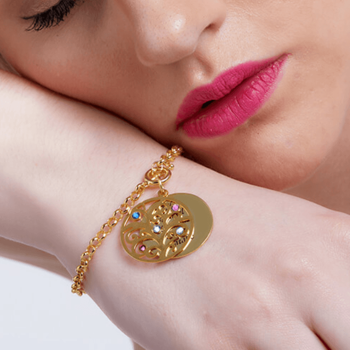 A woman wearing a gold charm bracelet with a tree pendant adorned with colored gemstones and a round disc, resting her head on her arm, with pink lips.