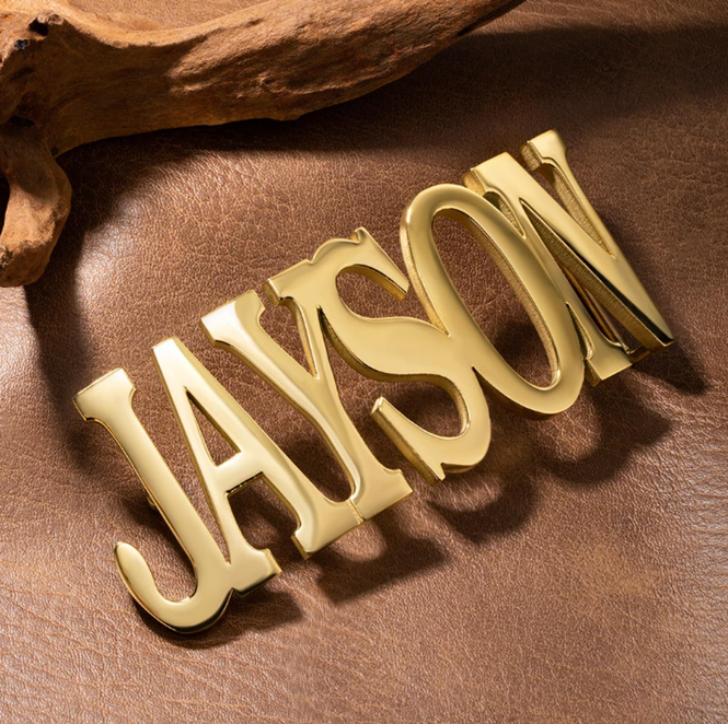 Shiny gold belt buckle with 'JAYSON' engraved, displayed on a textured brown leather surface with a rustic wood piece in the background.