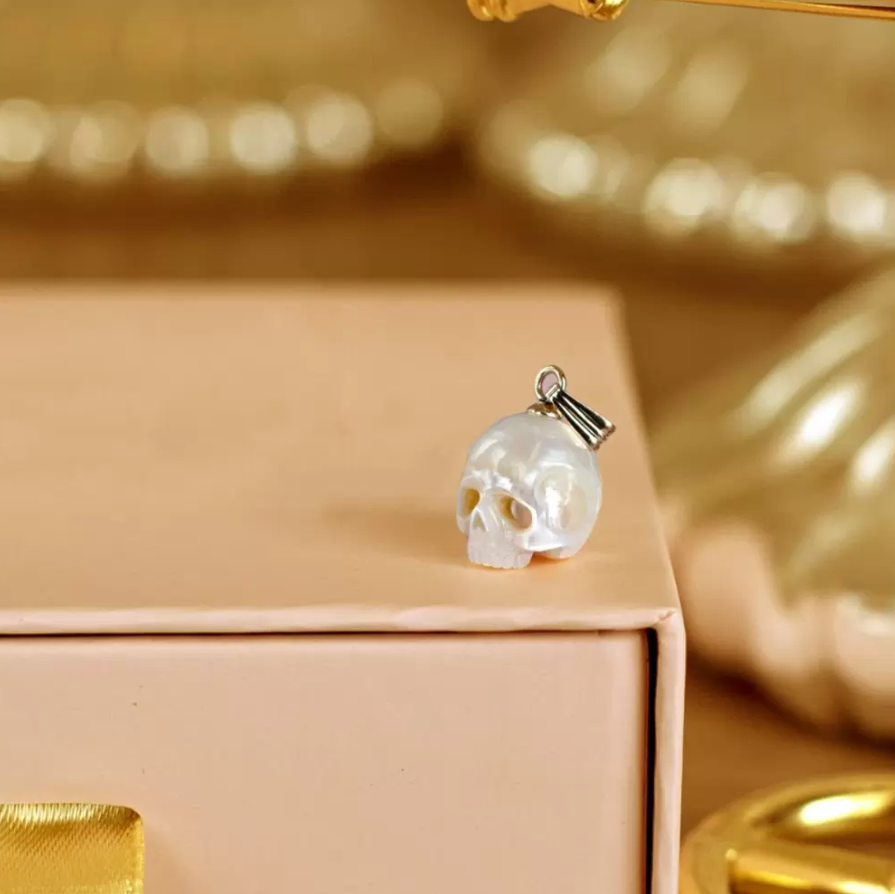 A luminous skull charm with a silver clasp set against a peach-toned box and golden bokeh background.