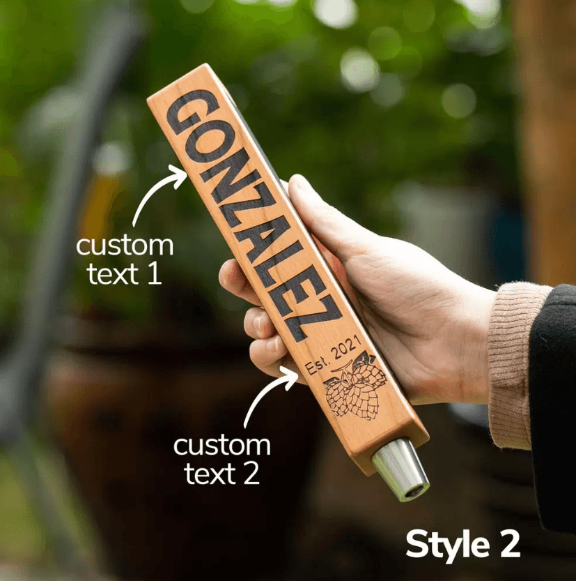 Hand holding a custom beer tap handle with personalized engraving, labeled "Gonzalez" with custom text areas and a hop graphic, in a garden setting.