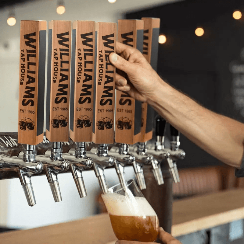 Custom beer tap handles with personalized engraving and chalkboard decals being used on a kegerator in a home bar, with a hand pouring a beer.