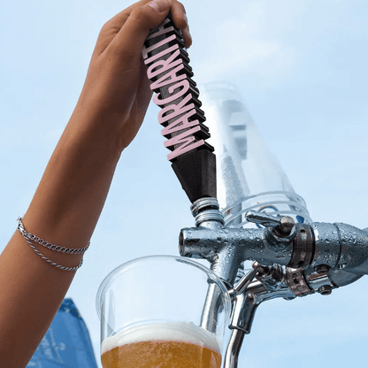 Custom 'MARGARITA' 3D printed beer tap handle in pink, attached to a silver beer dispenser, pouring into a glass, against a blue sky background.