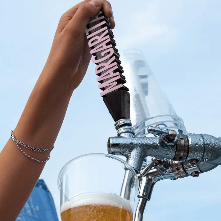 Custom 'MARGARITA' 3D printed beer tap handle in pink, attached to a silver beer dispenser, pouring into a glass, against a blue sky background.