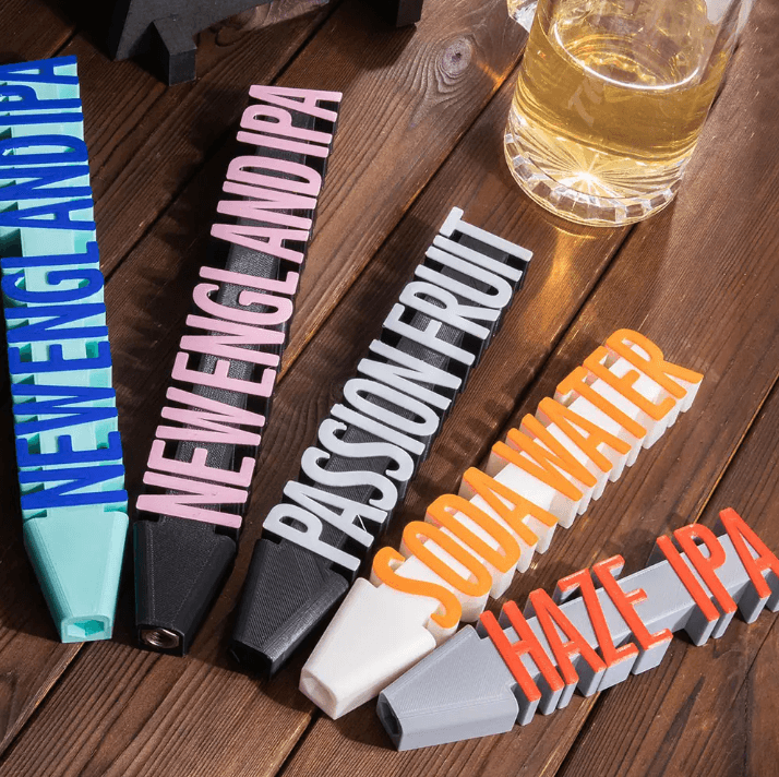 Four 3D printed beer tap handles with custom names: New England IPA, Passionfruit, Soda Water, Haze IPA, on a wooden surface next to a glass of beer.
