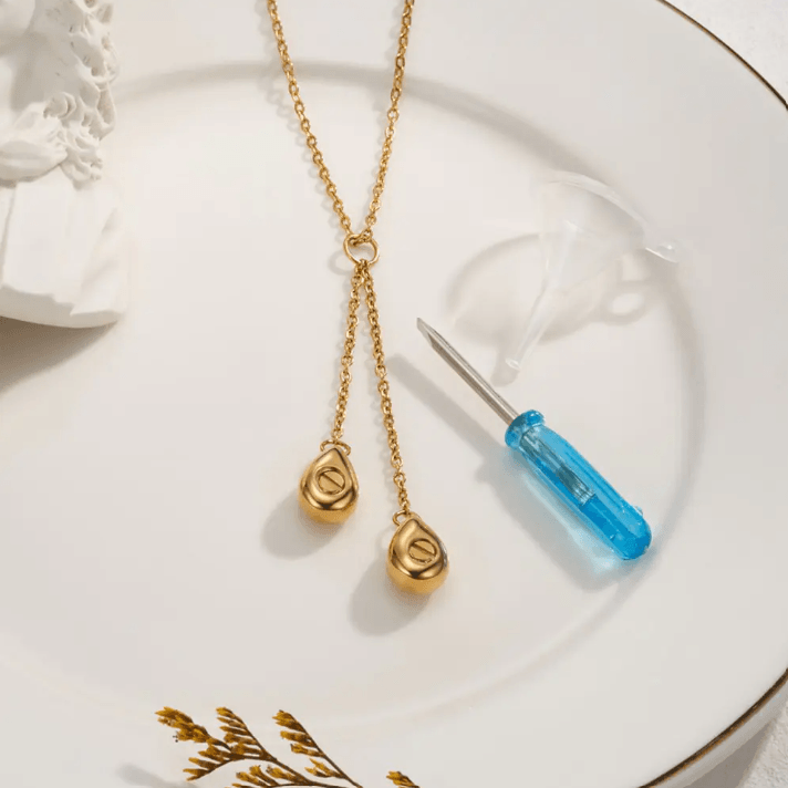 Gold necklace with two urn pendants displayed with a filling kit, including a funnel and screwdriver, on a white plate.