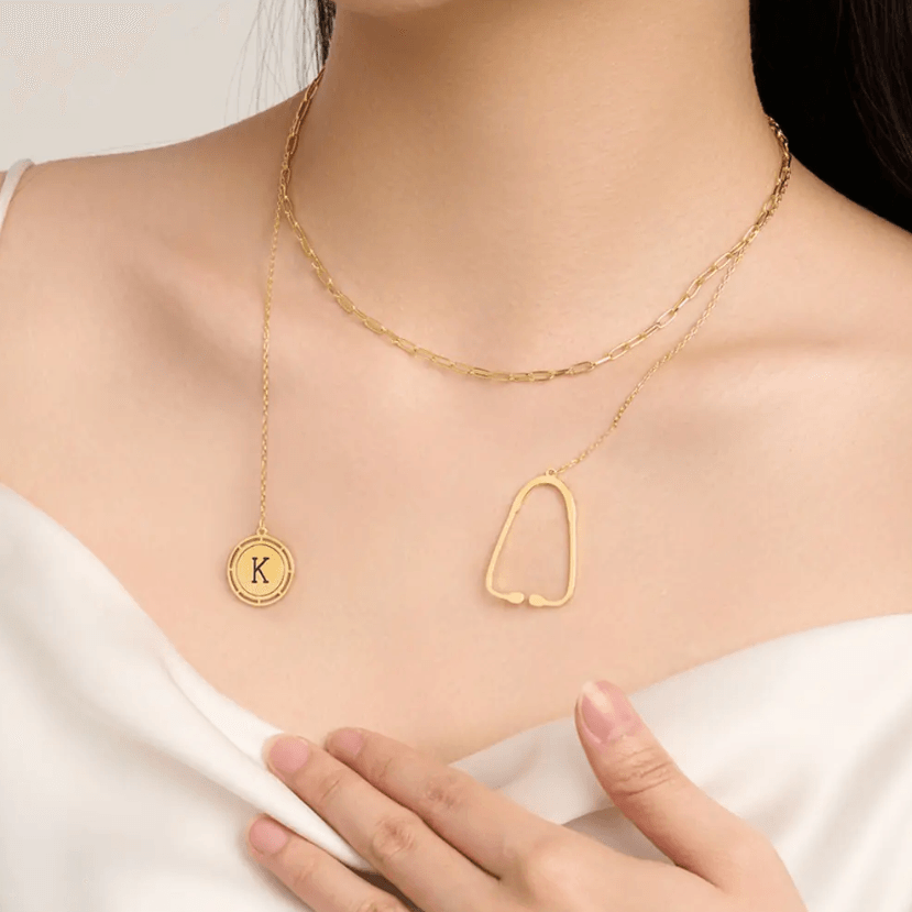 Layered gold necklace with a stethoscope pendant and a custom initial charm worn by a woman in a white top, hand resting near the charm.