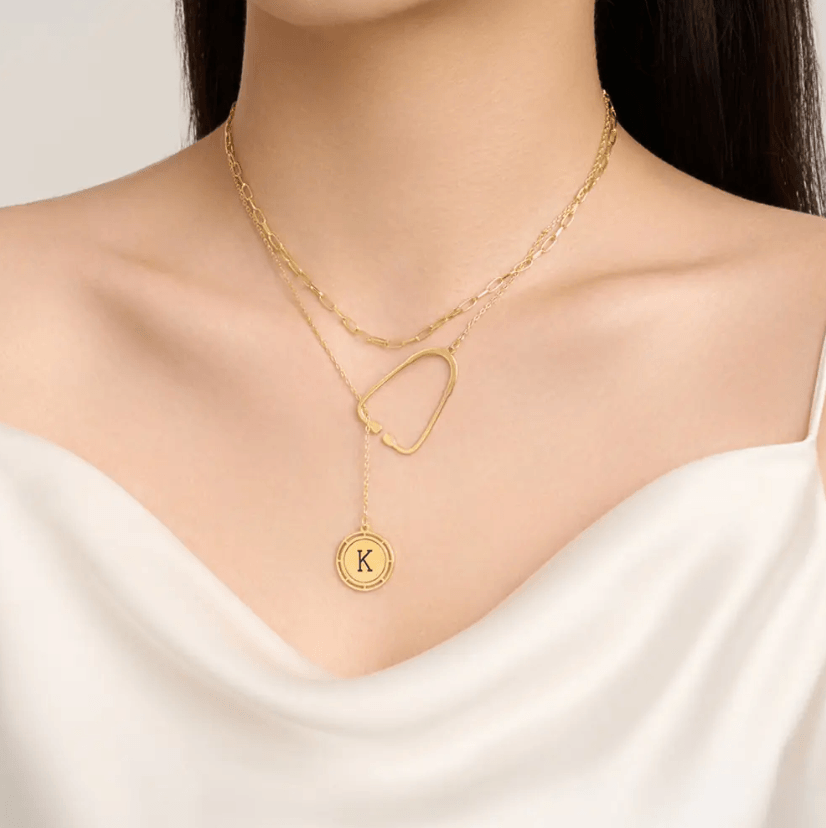 Layered gold necklace featuring a stethoscope pendant and a custom initial charm worn by a woman in a white top.