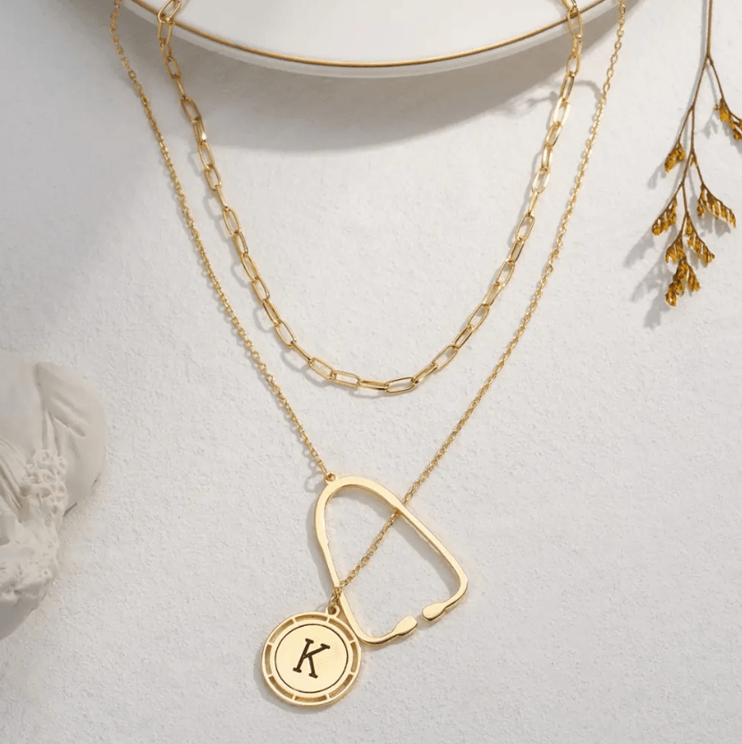 Layered gold necklace with a stethoscope pendant and a custom initial charm, displayed on a white background with decorative elements.