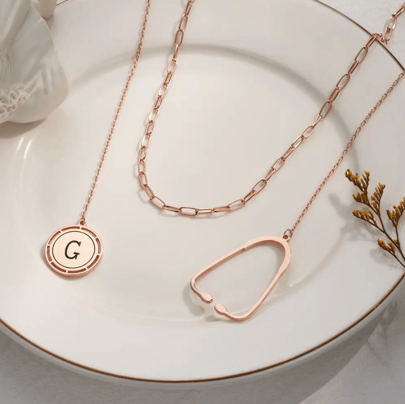 Layered rose gold necklace with a stethoscope pendant and a custom initial charm, displayed on a white plate with decorative elements.