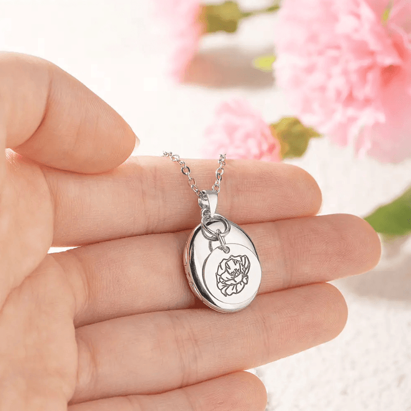 Hand holding a Personalized Birth Flower Necklace with an Oval Photo Locket in Sterling Silver, featuring a delicate flower engraving, against a pink flower background.