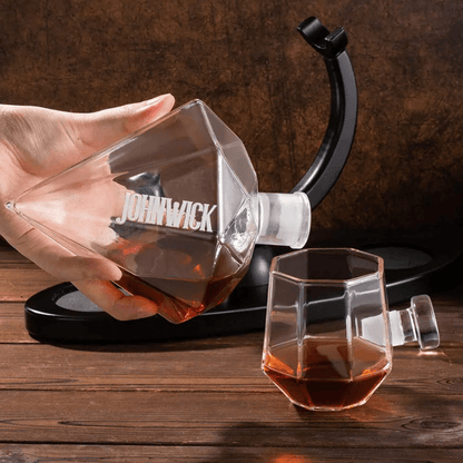 Hand pouring whiskey from a custom engraved diamond-shaped decanter labeled 'John Wick' into a geometric glass on a wooden table.