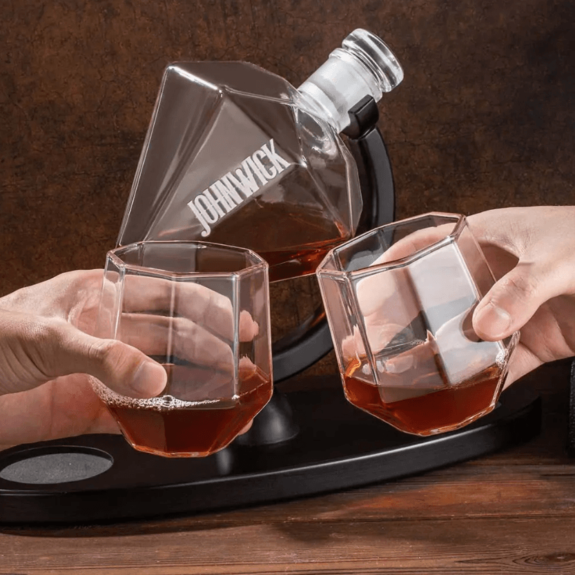 Two hands toasting with geometric glasses filled from a personalized diamond-shaped whiskey decanter labeled 'John Wick' on a wooden table.