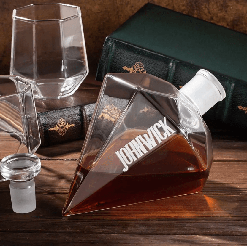 Personalized diamond-shaped whiskey decanter labeled 'John Wick' with two geometric glasses, resting on a wooden table near vintage books.