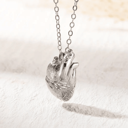 Silver necklace featuring a detailed, anatomical heart pendant on a chain, displayed on a textured white surface.