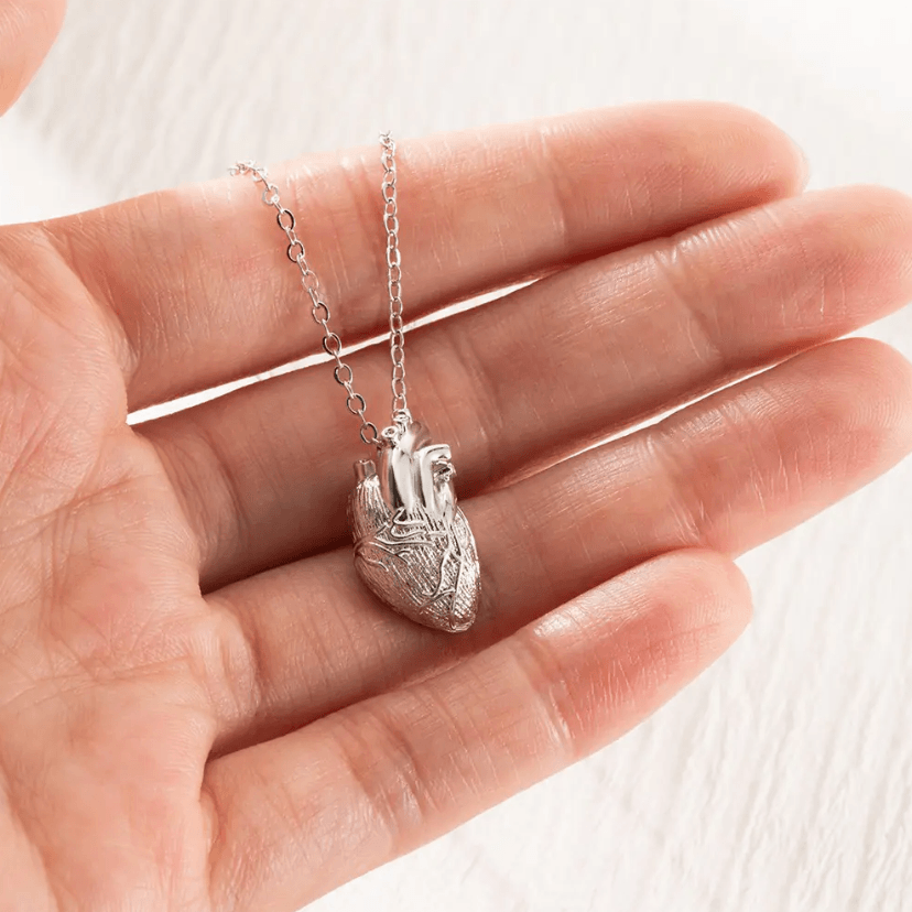 Silver anatomical heart pendant on a chain, held gently in a person's palm against a white background.