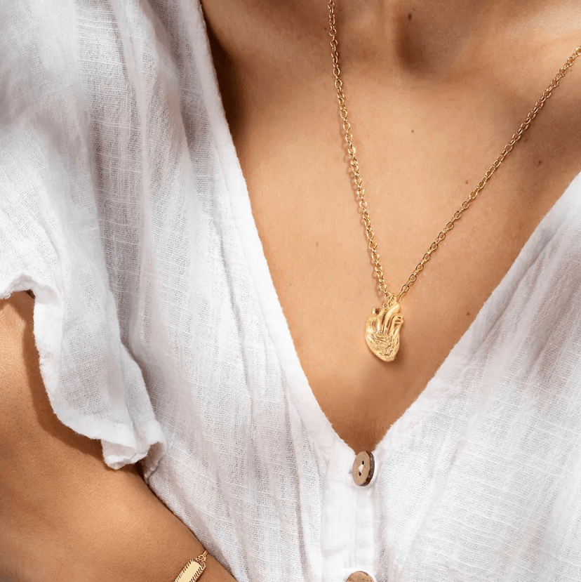 Gold anatomical heart necklace worn on a person's neck, contrasted against a white, textured blouse.