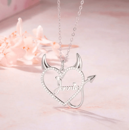 Silver necklace with a heart-shaped pendant featuring devil horns and tail, and the name "Jennie" in the center, surrounded by sparkling stones, displayed on a soft pink surface.