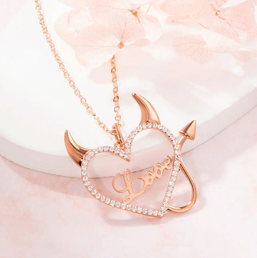 Rose gold necklace with a heart-shaped pendant featuring devil horns and tail, and the word "Love" in the center, surrounded by sparkling stones, displayed on a soft pink surface.