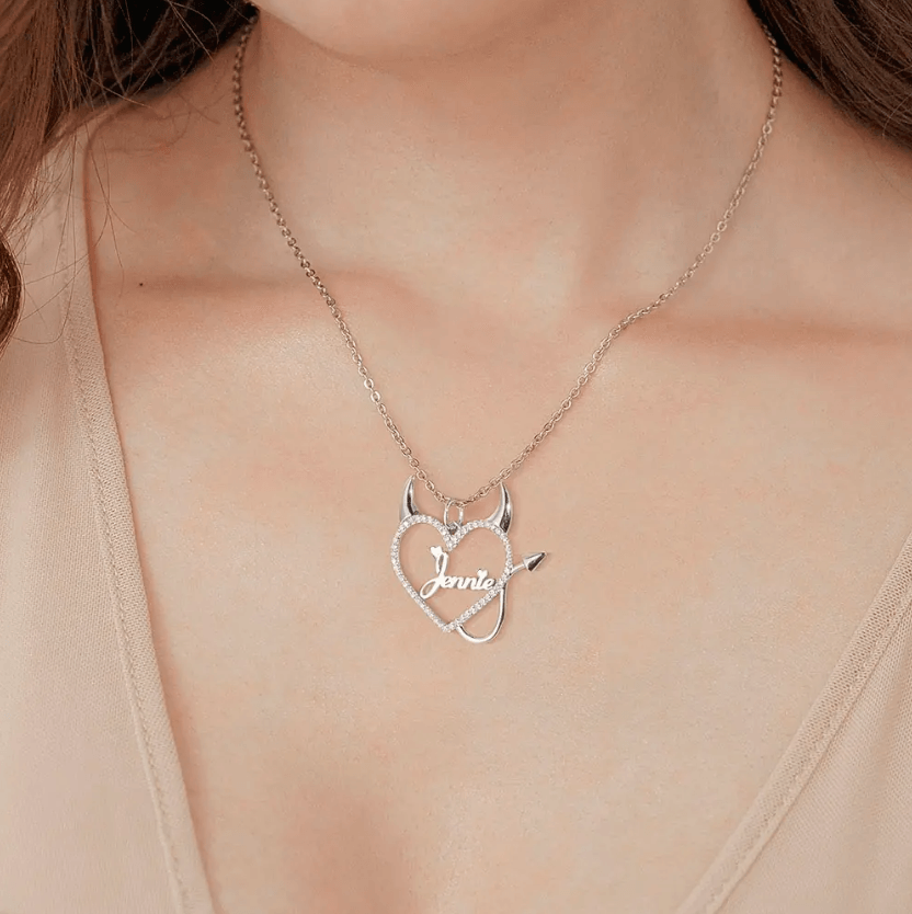 Woman wearing a silver necklace with a heart-shaped pendant featuring devil horns and tail, and the name "Jennie" in the center, surrounded by sparkling stones.