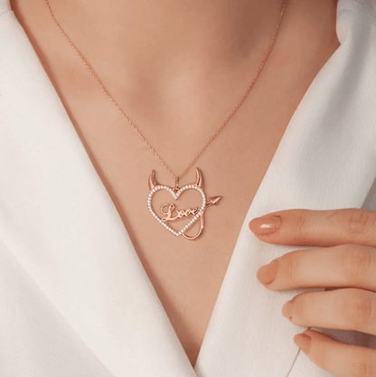 Woman wearing a rose gold necklace with a heart-shaped pendant featuring devil horns and tail, and the word "Love" in the center, surrounded by sparkling stones.