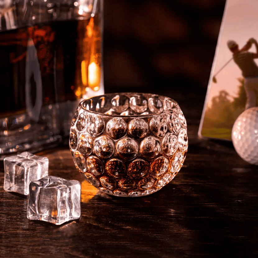 Golf ball-shaped whiskey glass filled with whiskey, surrounded by ice cubes, a whiskey decanter, and a golfing photo on a wooden table.