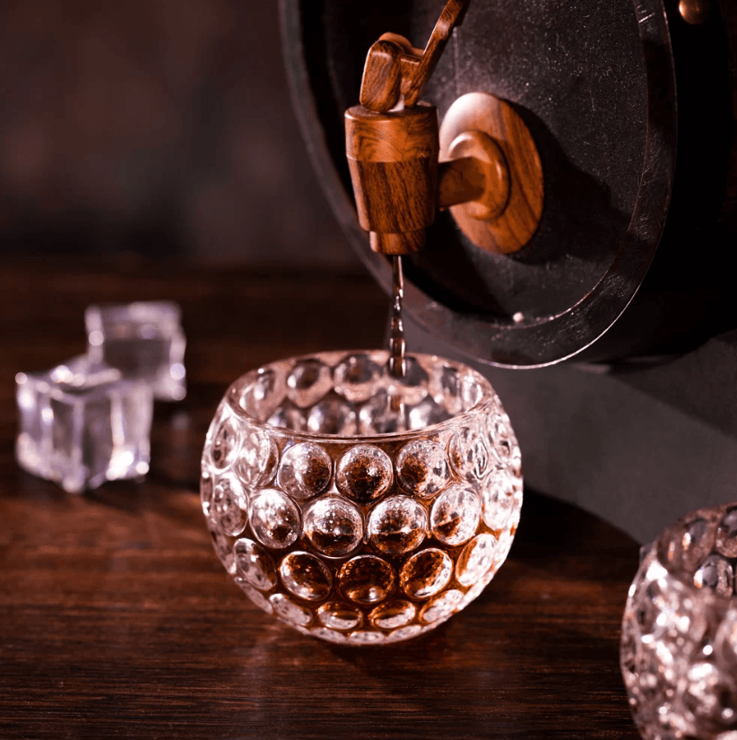 Whiskey being poured from a wooden barrel tap into a golf ball-shaped glass, with ice cubes on a wooden table.
