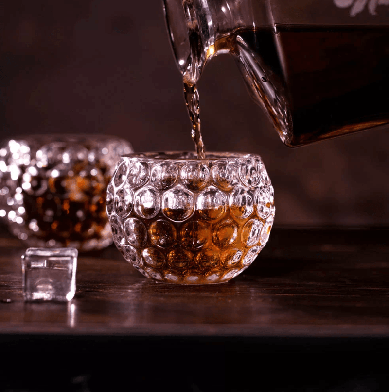 Whiskey being poured from a decanter into a golf ball-shaped glass, with another filled glass and ice cubes on a wooden table.