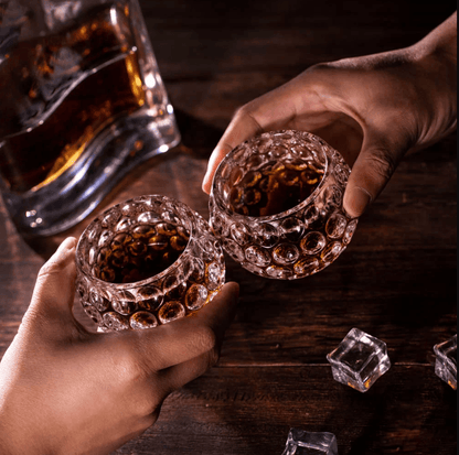Two hands holding golf ball-shaped whiskey glasses in a toast, with a whiskey decanter and ice cubes on a wooden table.