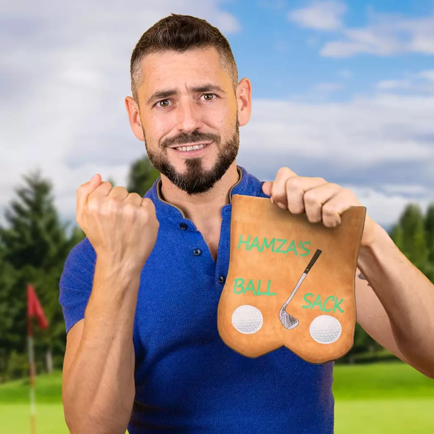Joyful bearded man in a blue polo shirt displaying a 'Hamza's Ball Sack' golf bag with a golf course in the background, ideal for a personalized golf gift.