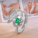 Silver memorial ring with angel wings design and a green heart-shaped stone nestled between baby feet, with the inscription "Forever in my heart" and a background of photos showing heart-shaped hands on a pregnant belly and baby feet.