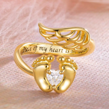 A gold-toned memorial ring with a heart-shaped stone cradled by feet, angel wings on top, and an inscription "Piece of my heart is in heaven".