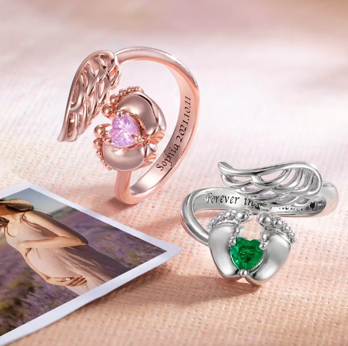 Two memorial rings with angel wings and heart-shaped stones cradled by feet; one rose gold with pink stone and "Sophia 02.10.2021" engraving, one silver with green stone and "Forever in my heart" inscription, beside a photo of a pregnant woman.