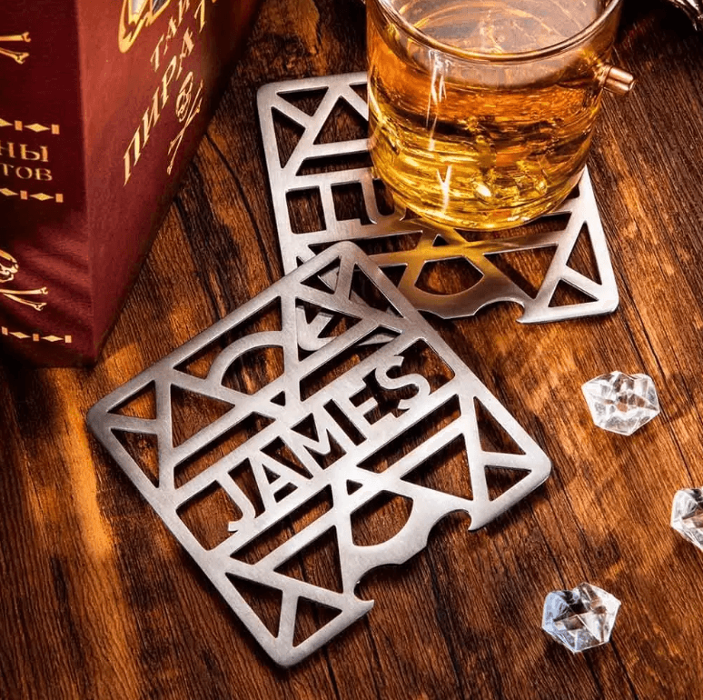 Personalized stainless steel coaster with 'JAMES' cutout, featuring a glass of whiskey and a book on a wooden table.