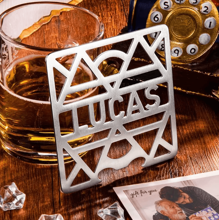 Custom 'LUCAS' stainless steel coaster near a whiskey glass, vintage phone, and gift card on a wooden table.