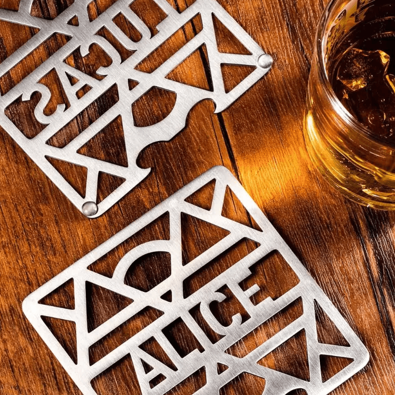 Two personalized stainless steel coasters, 'ALICE' and 'Lucas,' beside a glass of whiskey on a wooden table.